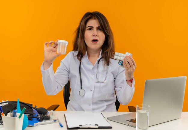 Doctora de mediana edad confundida con bata médica con estetoscopio sentado en el escritorio, trabajo en una computadora portátil con herramientas médicas que sostienen lata vacía y píldoras en una pared naranja aislada