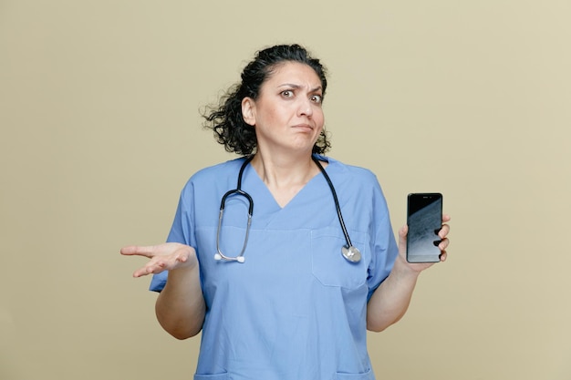 Doctora de mediana edad despistada con uniforme y estetoscopio alrededor del cuello que muestra el teléfono móvil y la mano vacía mirando la cámara aislada en el fondo verde oliva