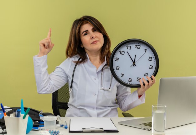 Doctora de mediana edad disgustada con bata médica y un estetoscopio sentado en el escritorio con herramientas médicas y una computadora portátil con reloj apuntando hacia arriba aislado