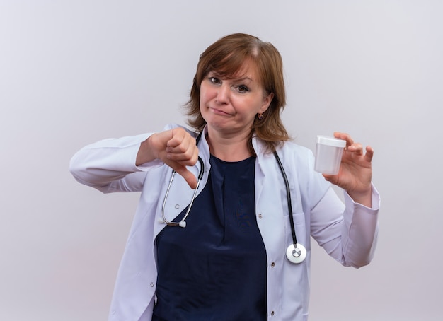 Doctora de mediana edad disgustada con bata médica y estetoscopio sosteniendo un vaso de precipitados médico y mostrando el pulgar hacia abajo sobre fondo blanco aislado