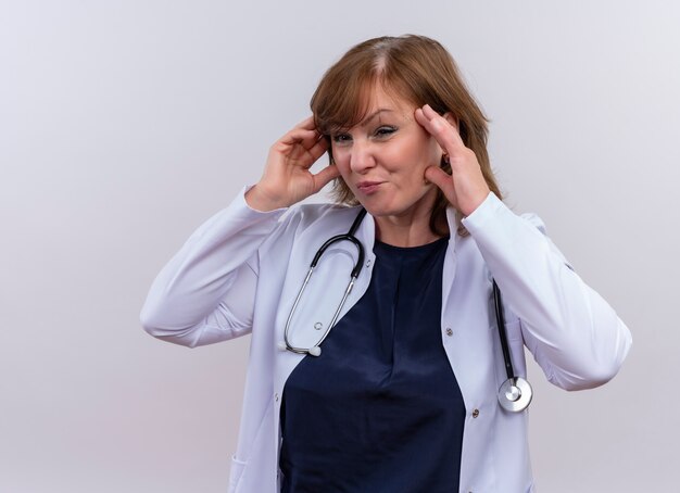 Doctora de mediana edad disgustada vistiendo bata médica y estetoscopio poniendo las manos en las sienes sobre fondo blanco aislado con espacio de copia