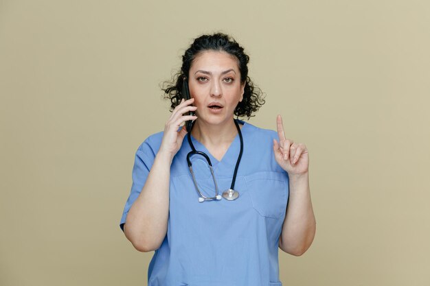 Doctora de mediana edad impresionada con uniforme y estetoscopio alrededor del cuello mirando a la cámara apuntando hacia arriba mientras habla por teléfono aislada en un fondo verde oliva