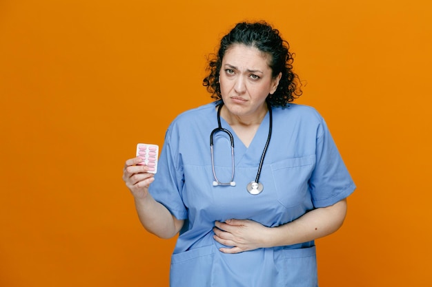 Doctora de mediana edad malsana con uniforme y estetoscopio alrededor de su cuello mostrando un paquete de cápsulas mirando a la cámara manteniendo la mano en el vientre aislado en un fondo naranja