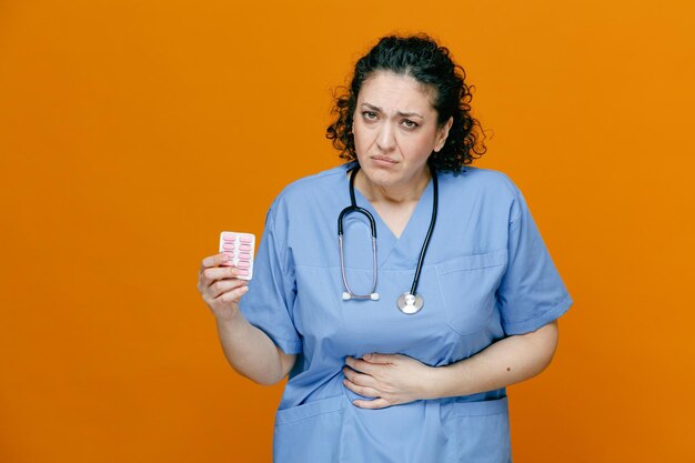 Doctora de mediana edad malsana con uniforme y estetoscopio alrededor de su cuello mostrando un paquete de cápsulas mirando a la cámara manteniendo la mano en el vientre aislado en un fondo naranja