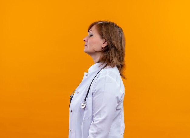 Doctora de mediana edad mirando seriamente con bata médica y estetoscopio de pie en la vista de perfil en la pared naranja aislada con espacio de copia