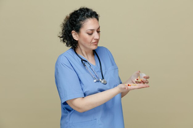 Doctora de mediana edad pensativa con uniforme y estetoscopio alrededor del cuello sosteniendo un recipiente de medición con pastillas y pastillas en la mano mirando pastillas aisladas en un fondo verde oliva