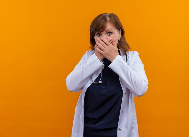 Doctora de mediana edad sorprendida con bata médica y estetoscopio poniendo las manos en la boca en la pared naranja aislada con espacio de copia