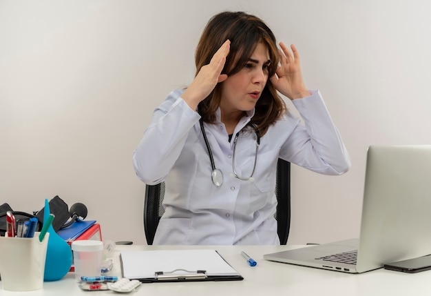 Doctora de mediana edad sorprendida con bata médica y estetoscopio sentado en el escritorio con portapapeles de herramientas médicas mirando portátil tocando la cabeza aislada