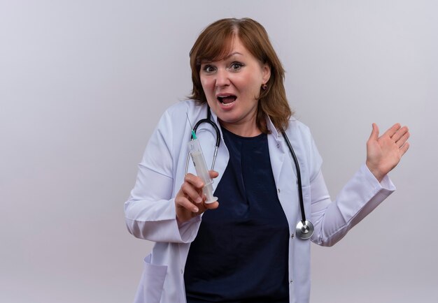 Doctora de mediana edad sorprendida vistiendo bata médica y estetoscopio con jeringa en pared blanca aislada con espacio de copia