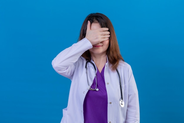 Doctora de mediana edad vistiendo bata blanca y con estetoscopio cubriendo los ojos con la mano sonriendo de pie sobre fondo azul.