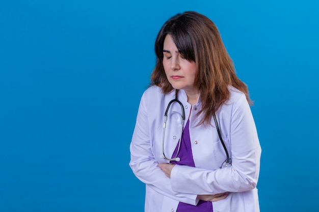 Doctora de mediana edad vistiendo bata blanca y con estetoscopio mirando malestar tocando el estómago que sufre de dolor de pie sobre fondo azul.