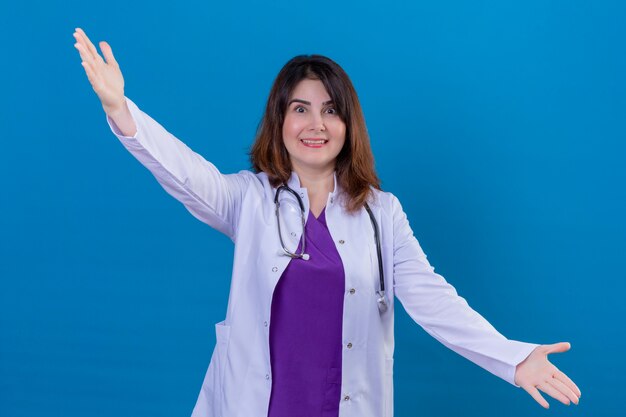Doctora de mediana edad vistiendo bata blanca y con estetoscopio sonriendo haciendo un gesto de bienvenida con las manos levantadas sobre fondo azul.