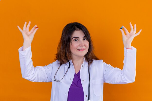 Doctora de mediana edad vistiendo bata blanca y con estetoscopio sosteniendo pastillas en las manos mirando a la cámara sonriendo de pie sobre fondo naranja