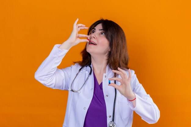 Doctora de mediana edad vistiendo bata blanca y con estetoscopio sosteniendo pastillas en las manos tratando de tomar una pastilla de pie sobre fondo naranja