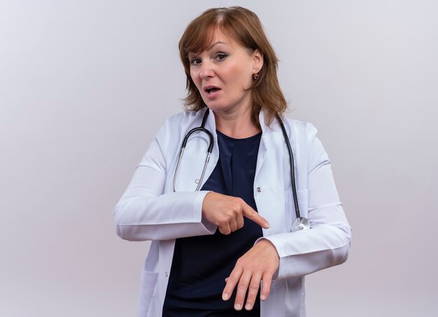 Doctora de mediana edad vistiendo bata médica y estetoscopio apuntando con el dedo a su mano en la pared blanca aislada con espacio de copia