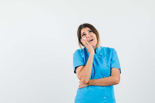 Doctora mirando hacia arriba, apoyando la mejilla en la mano en uniforme azul y mirando soñadora