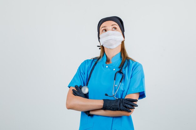 Doctora mirando hacia arriba con los brazos cruzados en uniforme, máscara, guantes