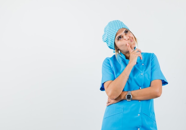 Doctora mirando hacia arriba con el dedo en la mejilla en uniforme azul y mirando vacilante