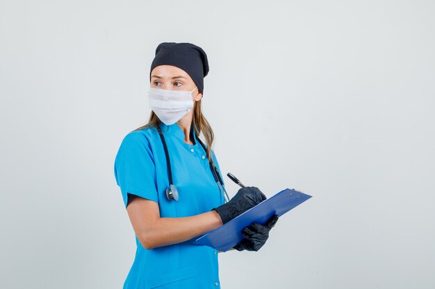 Doctora mirando hacia atrás mientras escribe en el portapapeles con uniforme, guantes, máscara y aspecto serio.