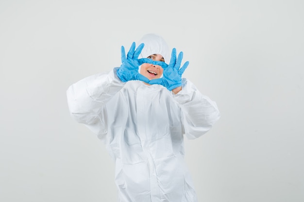 Doctora mostrando gesto de corazón en traje protector, guantes y mirando feliz.