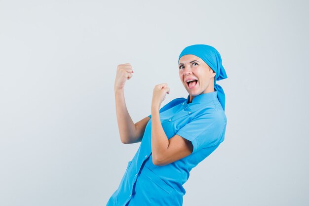 Doctora mostrando gesto ganador en uniforme azul y mirando feliz, vista frontal.