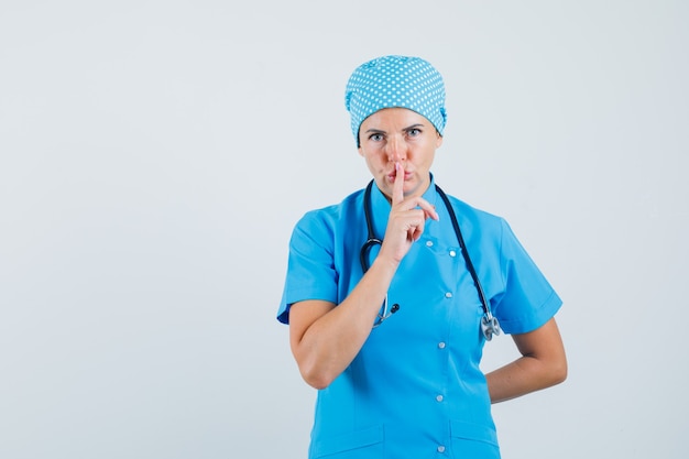 Doctora mostrando gesto de silencio en uniforme azul y mirando con cuidado. vista frontal.