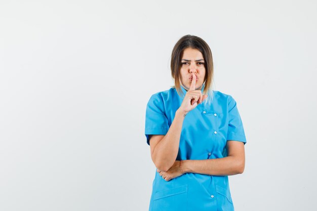 Doctora mostrando gesto de silencio en uniforme azul y mirando con cuidado