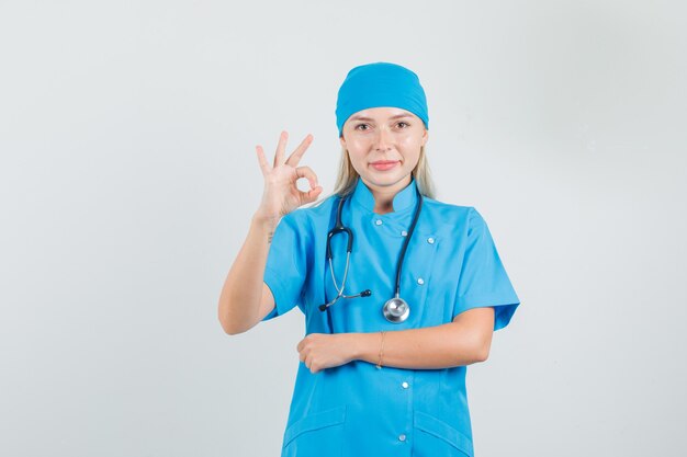 Doctora mostrando signo ok en uniforme azul y mirando complacido.