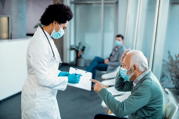 Doctora negra y anciano con máscaras faciales analizando informes médicos en un pasillo del hospital