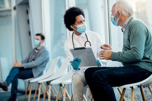 Doctora negra y anciano con máscaras faciales mientras hablan de un informe médico en el hospital