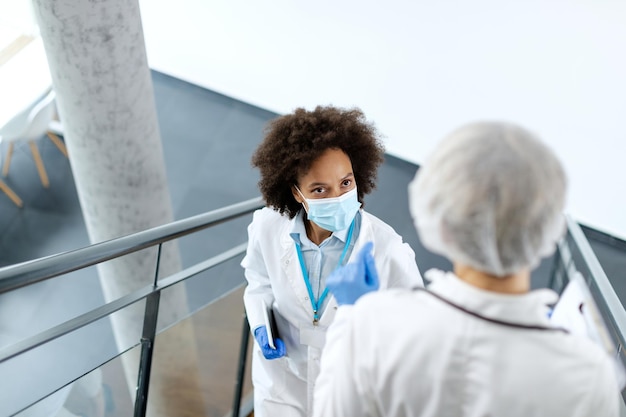 Foto gratuita doctora negra con mascarilla protectora mientras pasa junto a su colega en el pasillo del hospital