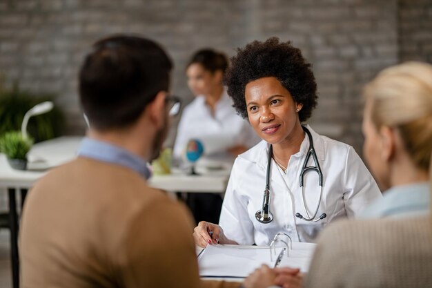 Una doctora negra sonriente que se comunica con una pareja mientras revisa su historial médico en la clínica