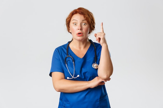 Doctora pelirroja abrumada, enfermera de mediana edad en bata tiene sugerencia, diciendo idea o plan, levantando el gesto de eureka del dedo índice con expresión preocupada, de pie
