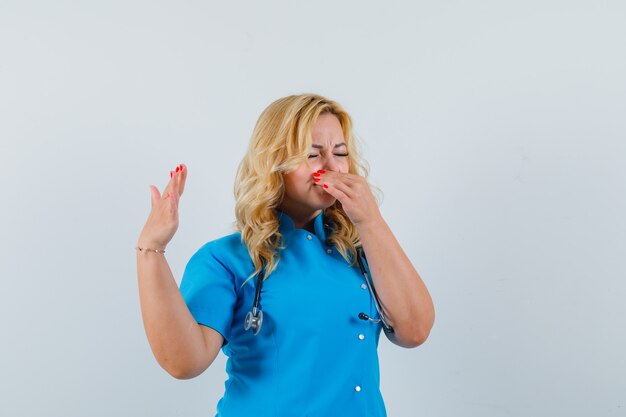 Doctora pellizcando su nariz en uniforme azul y mirando disgustada.