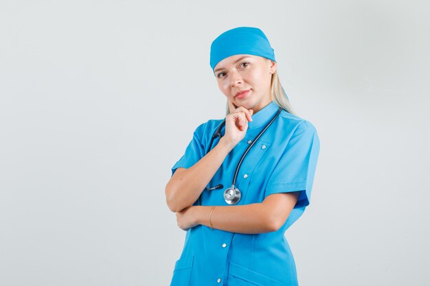 Doctora pensando con el dedo en la mejilla en uniforme azul y mirando esperanzada.