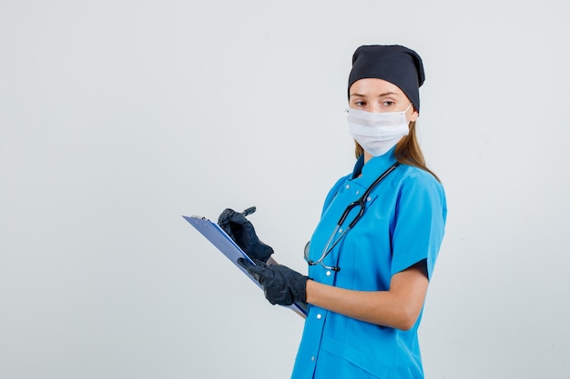 Doctora pensando mientras toma notas en el portapapeles en uniforme, guantes, máscara.