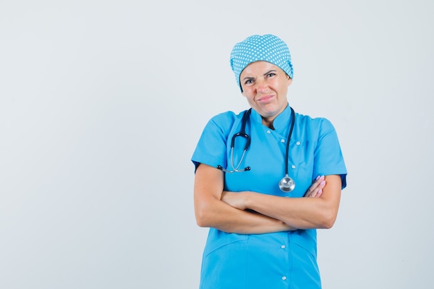 Doctora de pie con los brazos cruzados en uniforme azul y mirando insatisfecho. vista frontal.