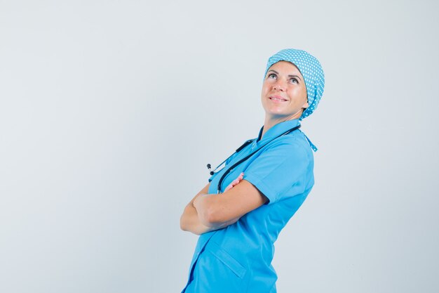 Doctora de pie con los brazos cruzados en uniforme azul y mirando pensativa. .