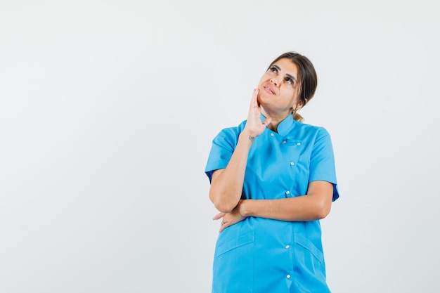 Doctora de pie en pose de pensamiento en uniforme azul y mirando soñadora