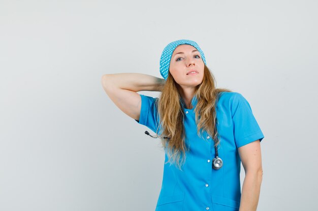 Doctora posando con la mano en el cuello en uniforme azul y mirando confiada.