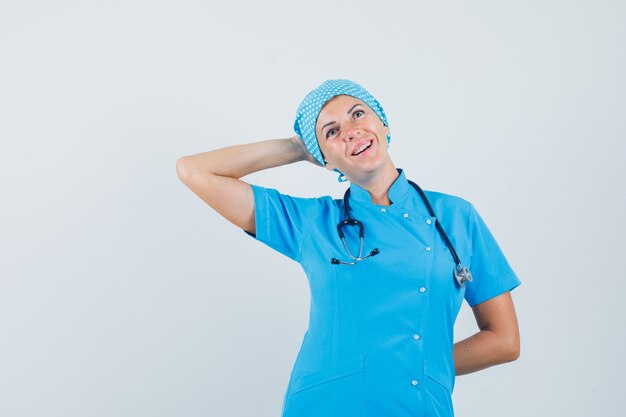 Doctora posando con la mano detrás de la cabeza en uniforme azul y mirando esperanzada. vista frontal.