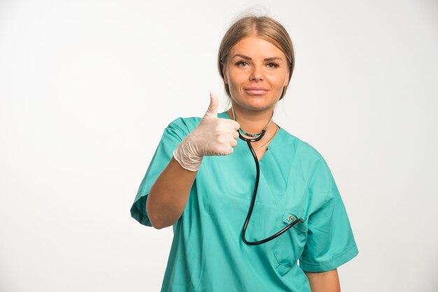 Doctora rubia en uniforme azul con estetoscopio en el cuello haciendo pulgar arriba signo.