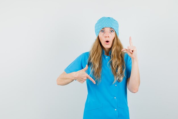 Doctora señalando con el dedo hacia arriba y hacia abajo en uniforme azul y mirando maravillada.