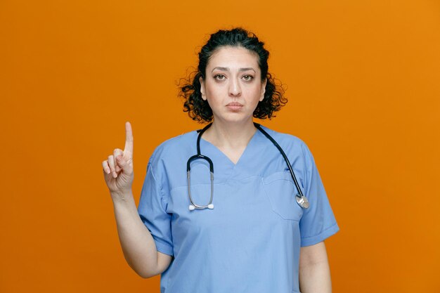 Una doctora seria de mediana edad con uniforme y estetoscopio alrededor del cuello mirando la cámara apuntando hacia arriba aislada en el fondo naranja