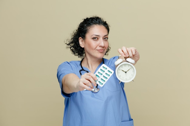 Doctora seria de mediana edad con uniforme y estetoscopio alrededor del cuello mirando la cámara estirando el despertador y el paquete de cápsulas hacia la cámara aislada en el fondo de oliva