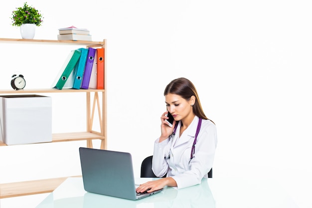 Doctora seria sentada en su escritorio mientras llama a alguien por teléfono y usa su computadora portátil