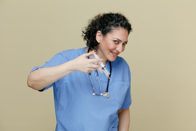 Foto gratuita una doctora sonriente de mediana edad con uniforme y estetoscopio alrededor del cuello sosteniendo una jeringa con una aguja en el aire mirando a la cámara simulando inyectarle a alguien aislado en un fondo verde oliva