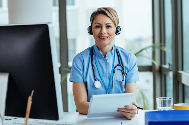 Una doctora sonriente que usa auriculares mientras trabaja en un centro de llamadas médicas y mira la cámara