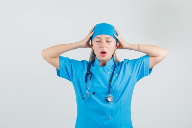 Doctora sosteniendo la cabeza con las manos en uniforme azul y mirando exhausto.