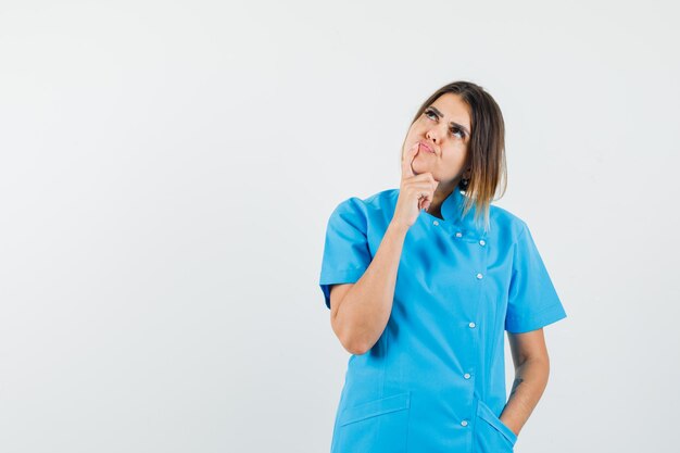 Doctora sosteniendo el dedo en la barbilla mientras mira hacia arriba en uniforme azul y mirando pensativo